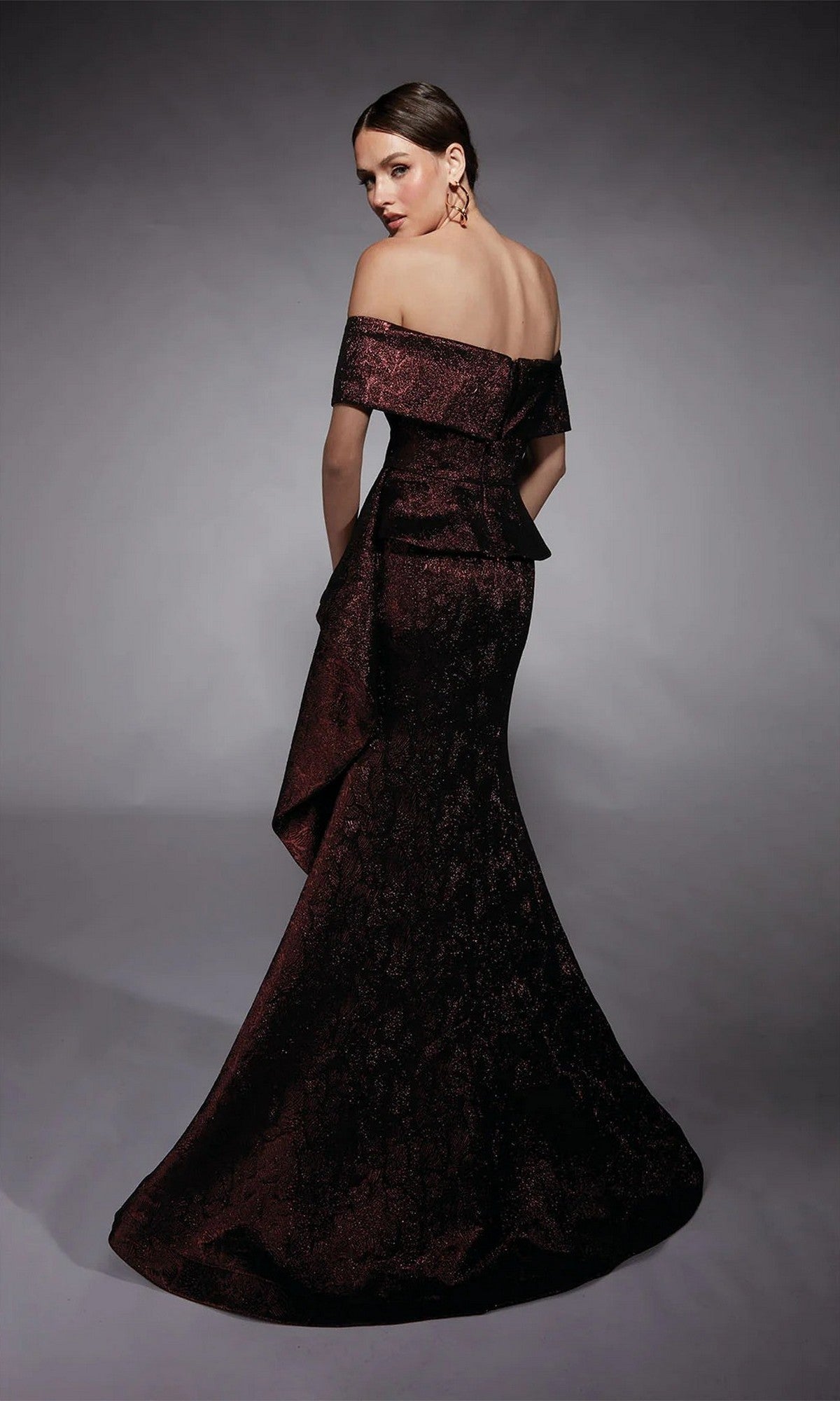 A woman in the Alyce 27707 long formal dress with an off-the-shoulder neckline and fit-and-flare jacquard design stands with her back to the camera against a gray background—an elegant choice for a mother-of-the-bride.